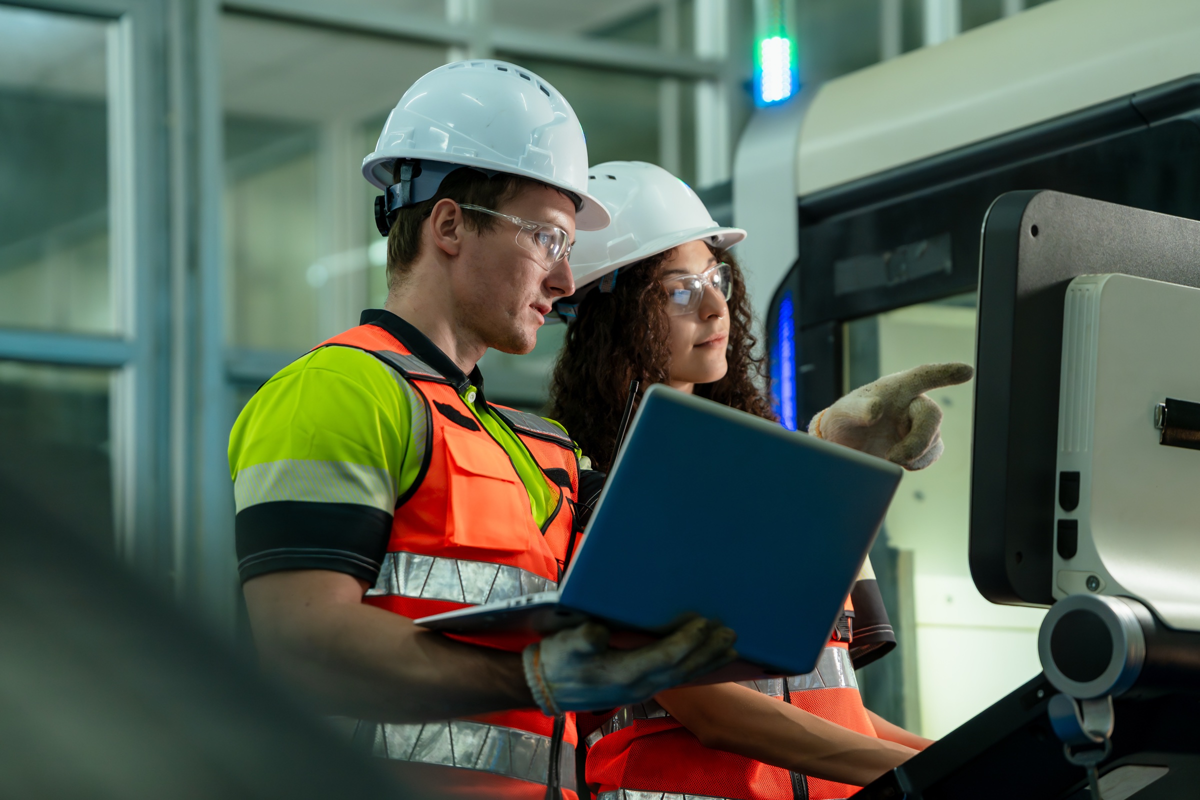 Two manufacturing employees wearing safety gear review equipment with a laptop.